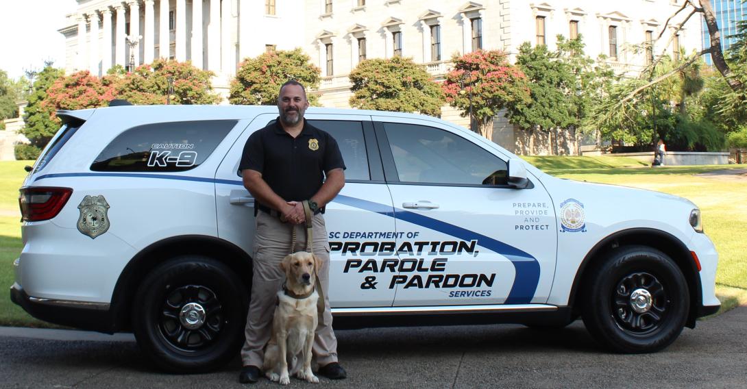 Agent Benjy and K-9 Chip standing in beside the SCDPPPS vehicle.